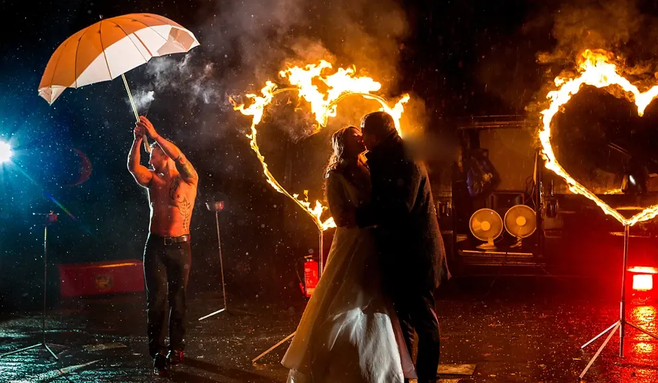 Brautpaar küsst sich zwischen zwei Flammenherzen im Regen, ein Feuerkünstler hält einen Schirm